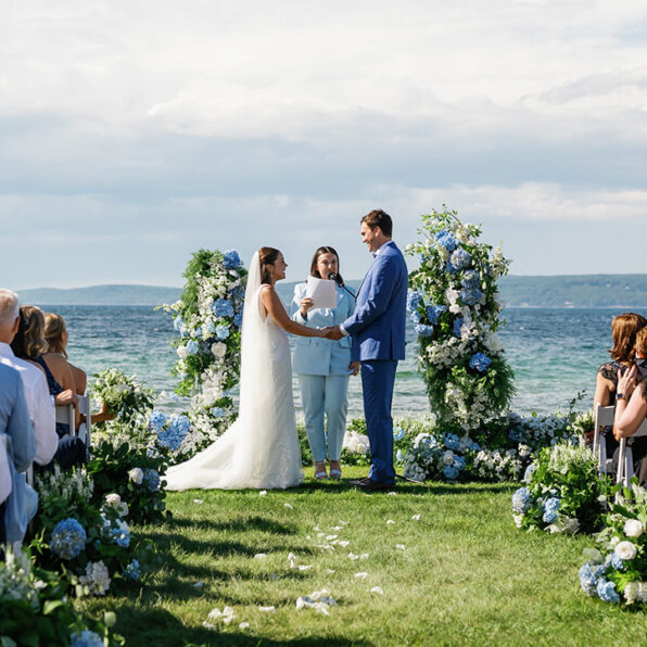 A bride and groom stand facing each other at an outdoor wedding ceremony by the sea. A person officiates, and guests are seated nearby. Blue floral decorations surround the couple.