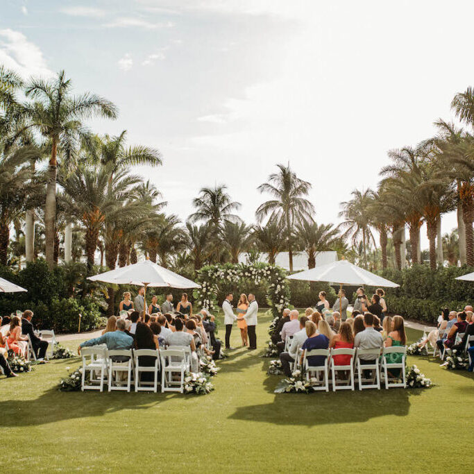 A wedding ceremony is taking place outdoors on a sunny day, with guests seated on white chairs arranged in a circular pattern around the couple. Palm trees and white umbrellas are in the background.