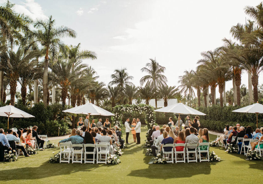 A wedding ceremony is taking place outdoors on a sunny day, with guests seated on white chairs arranged in a circular pattern around the couple. Palm trees and white umbrellas are in the background.