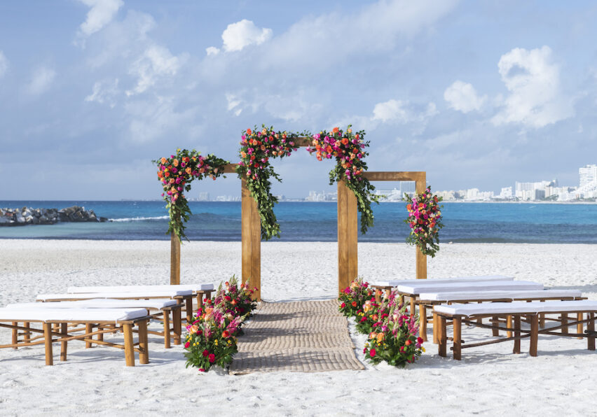 Wooden wedding arch decorated with colorful flowers on a sandy beach with benches, overlooking the ocean and Cancun city skyline in the distance.