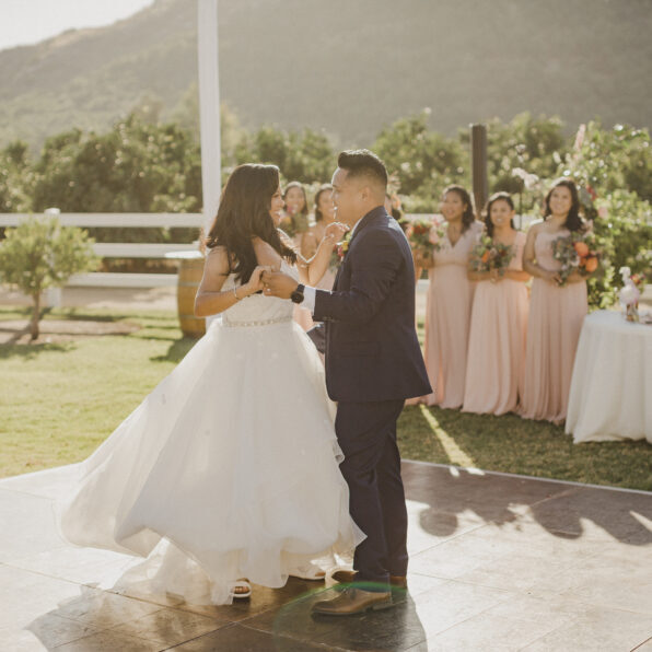 A bride and groom do their First Dance outdoors at their wedding reception, with bridesmaids in pink dresses watching in the background.