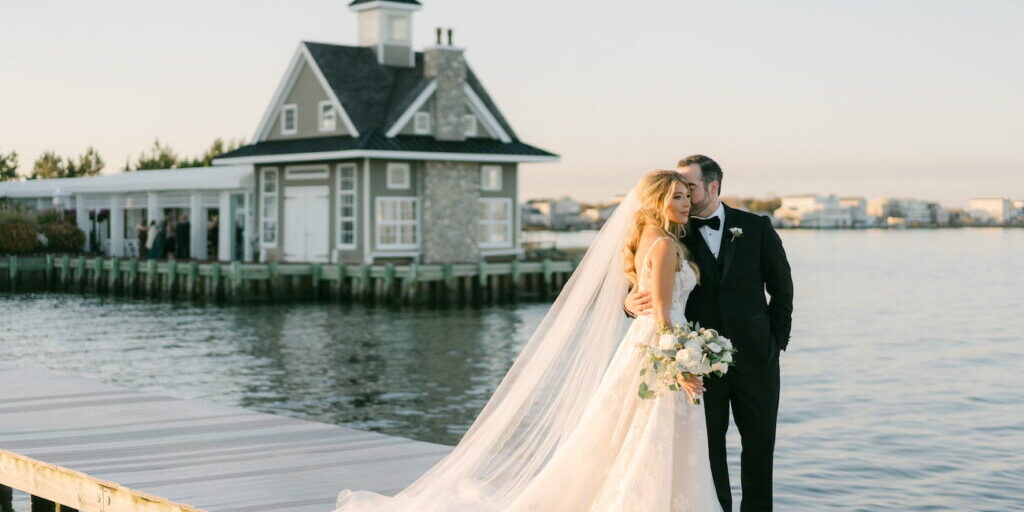 Bride and groom stand on a dock by the water, with the groom kissing the bride's forehead. A house on the shore is visible in the background.