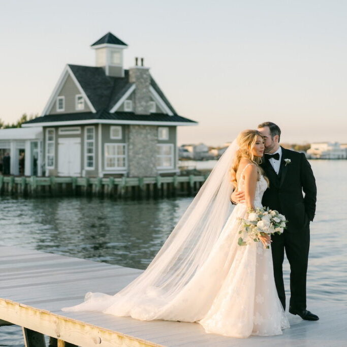 Bride and groom stand on a dock by the water, with the groom kissing the bride's forehead. A house on the shore is visible in the background.