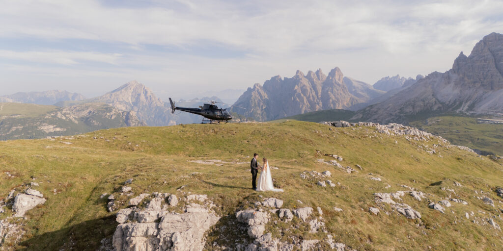 A bride and groom stand on a grassy mountaintop with a helicopter parked behind them and mountains in the background.
