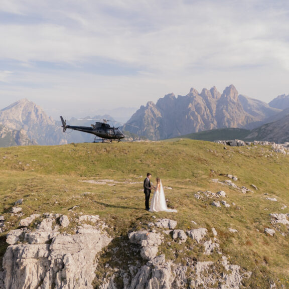A bride and groom stand on a grassy mountaintop with a helicopter parked behind them and mountains in the background.