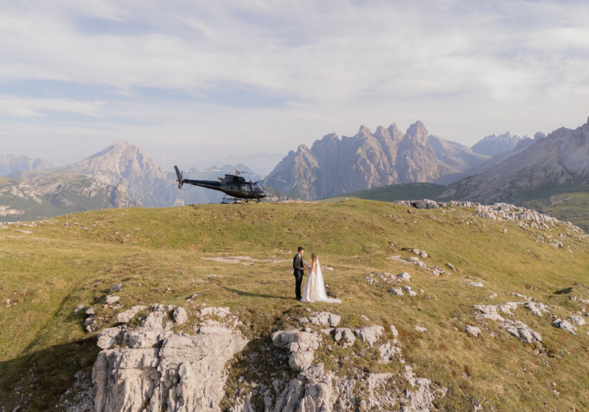 A bride and groom stand on a grassy mountaintop with a helicopter parked behind them and mountains in the background.