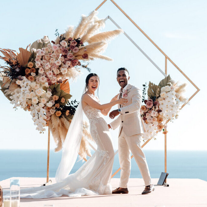 A couple stands at an altar adorned with flowers, exchanging vows under a canopy. Guests are seated on both sides, facing the ocean under a clear sky.