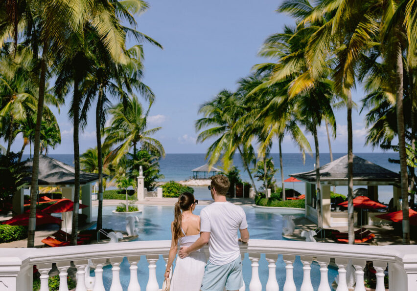 A couple standing together, overlooking Jewel Grande Montego Bay's luxurious pool surrounded by palm trees, facing the ocean.