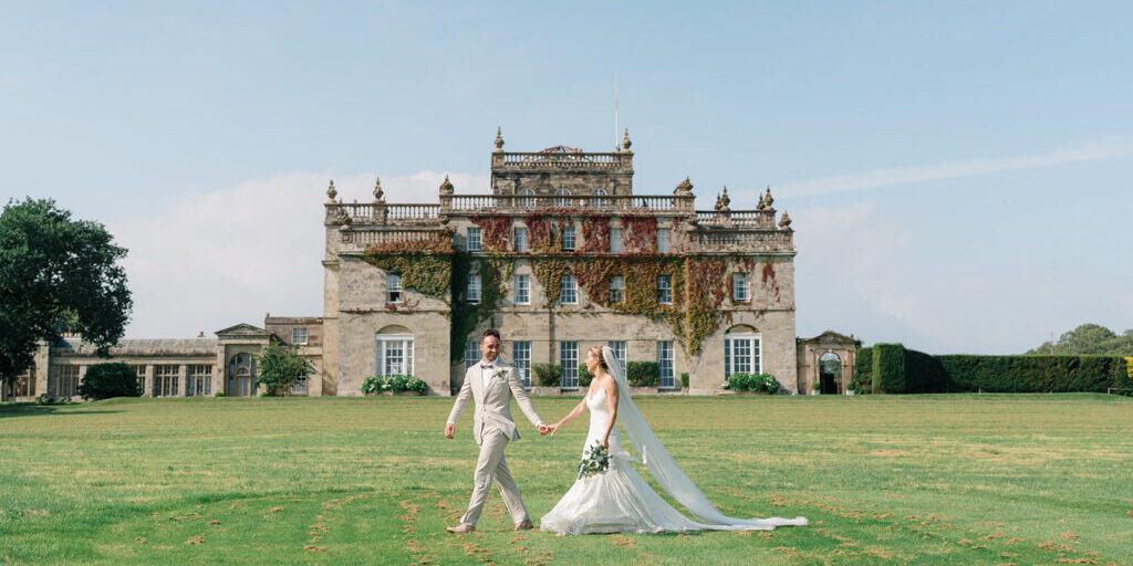A bride and groom walk hand in hand on a lawn in front of a large, historic stone mansion covered in ivy.