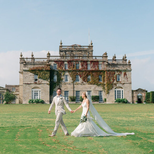 A bride and groom walk hand in hand on a lawn in front of a large, historic stone mansion covered in ivy.