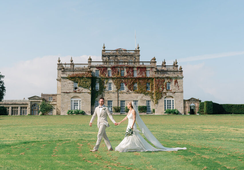 A bride and groom walk hand in hand on a lawn in front of a large, historic stone mansion covered in ivy.