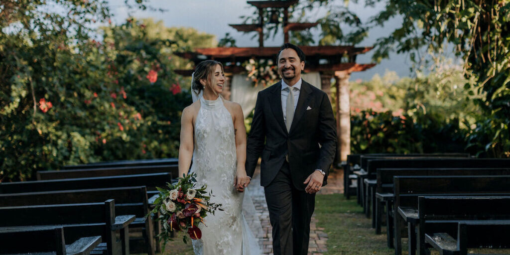 A bride in a white dress and a groom in a black suit walk hand in hand down an outdoor aisle, surrounded by greenery and empty benches.