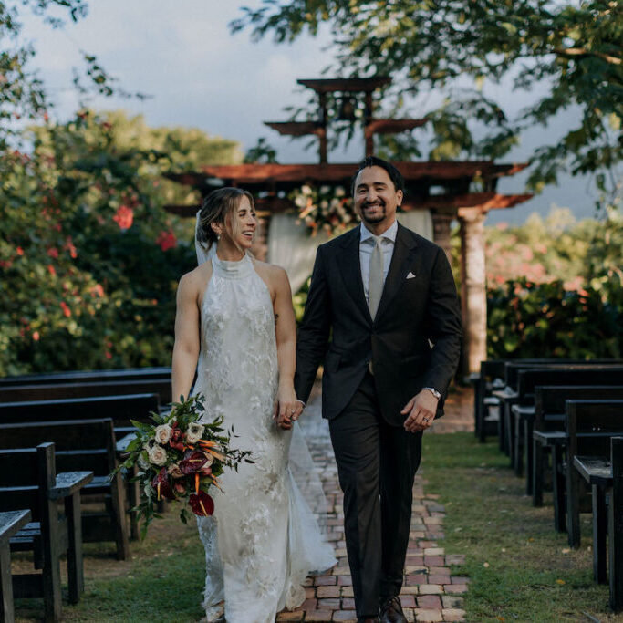 A bride in a white dress and a groom in a black suit walk hand in hand down an outdoor aisle, surrounded by greenery and empty benches.