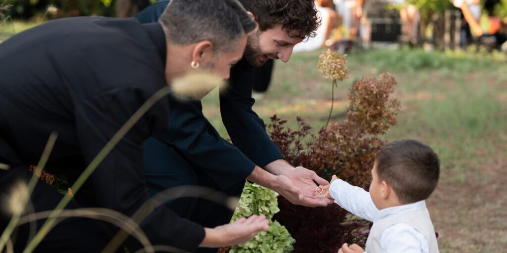 Two adults in dark clothing crouch and reach their hands toward a small child in white, who is standing outdoors among plants.