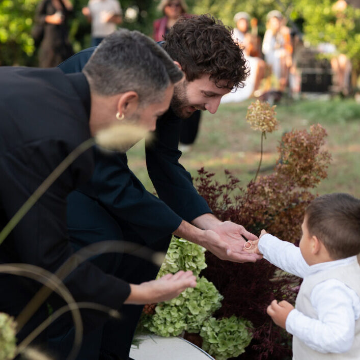 Two adults in dark clothing crouch and reach their hands toward a small child in white, who is standing outdoors among plants.