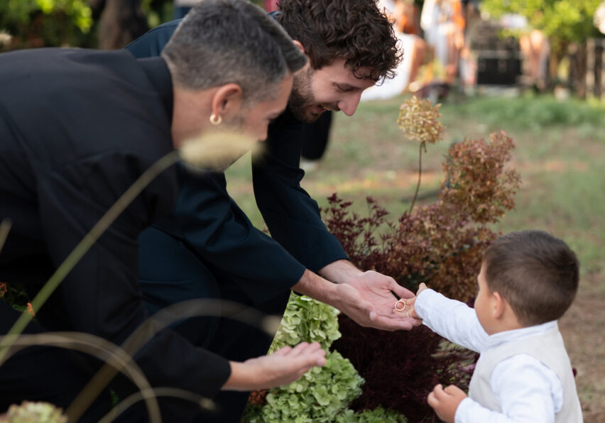 Two adults in dark clothing crouch and reach their hands toward a small child in white, who is standing outdoors among plants.
