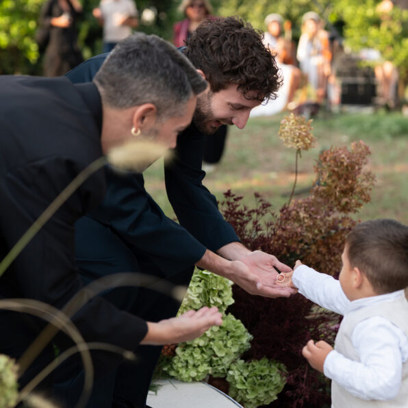 Two adults in dark clothing crouch and reach their hands toward a small child in white, who is standing outdoors among plants.