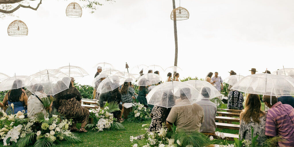 Guests sit on benches holding clear umbrellas at an outdoor wedding ceremony on a grassy lawn, surrounded by flowers and greenery.