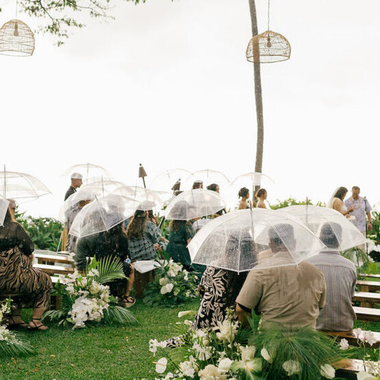 Guests sit on benches holding clear umbrellas at an outdoor wedding ceremony on a grassy lawn, surrounded by flowers and greenery.