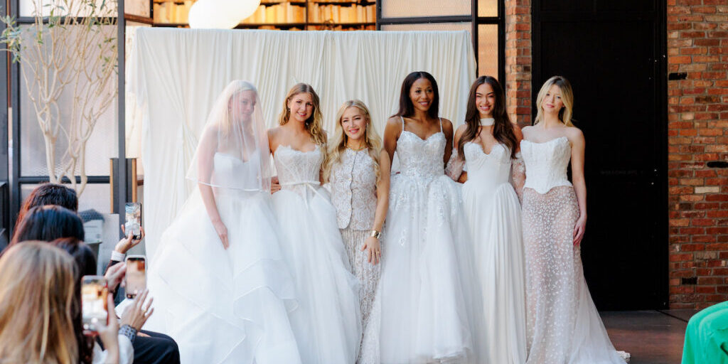 Six women wearing white bridal gowns pose together indoors at what appears to be a fashion event, with seated audience members visible in the foreground.