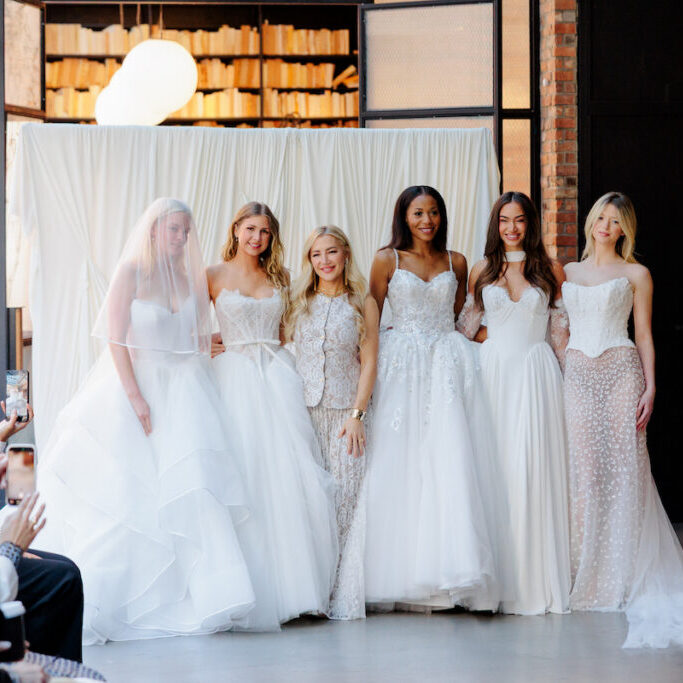Six women wearing white bridal gowns pose together indoors at what appears to be a fashion event, with seated audience members visible in the foreground.