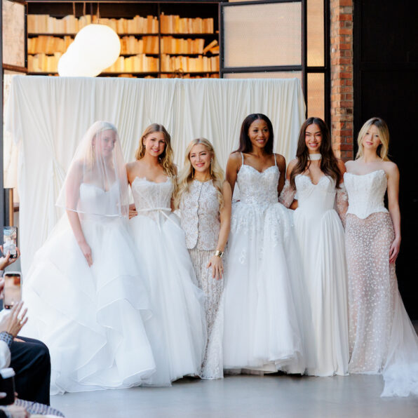 Six women wearing white bridal gowns pose together indoors at what appears to be a fashion event, with seated audience members visible in the foreground.