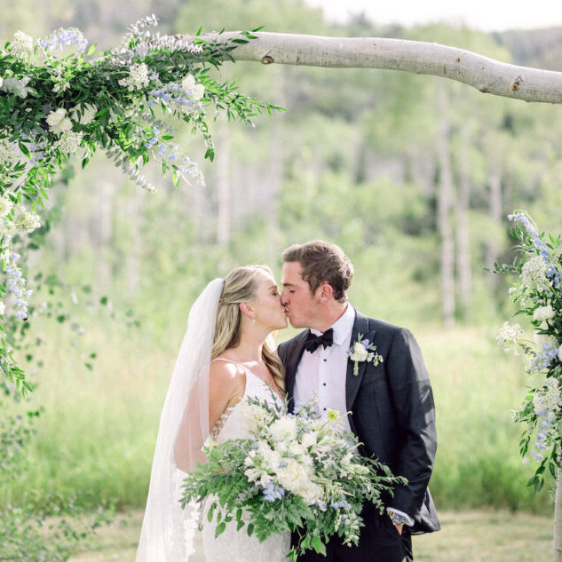 A bride and groom kiss under a floral arch in a lush, green meadow in Steamboat Springs, Colorado.