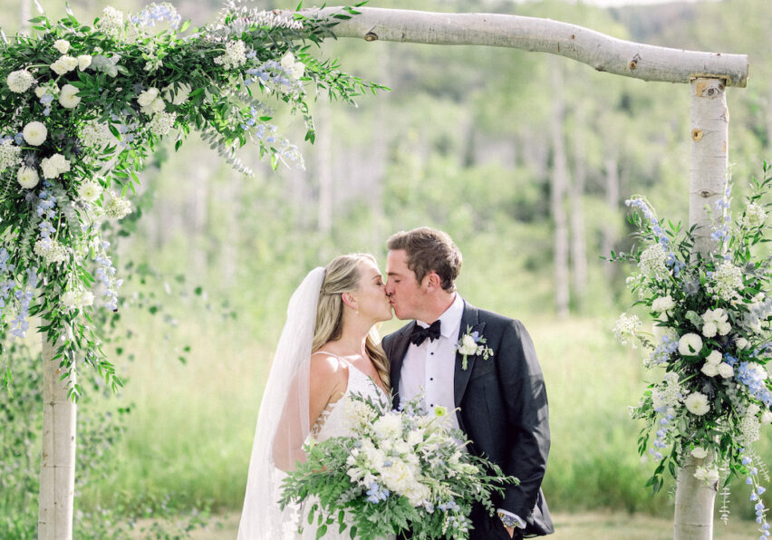 A bride and groom kiss under a floral arch in a lush, green meadow in Steamboat Springs, Colorado.