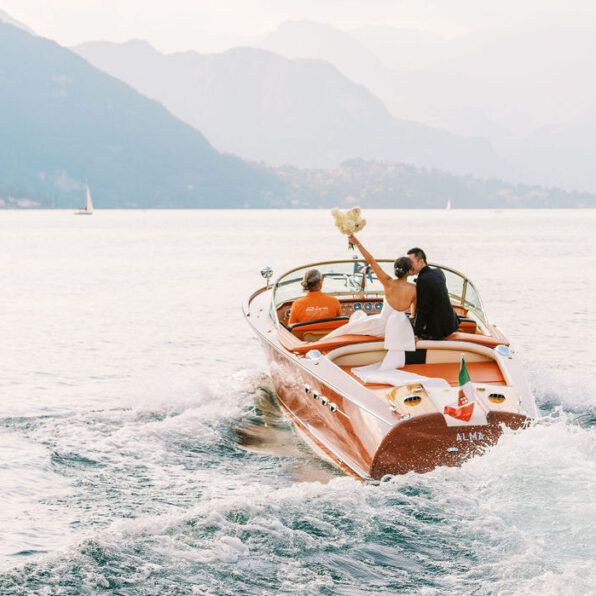 A couple dressed in wedding attire sits on the back of a wooden speedboat, with the bride holding a bouquet aloft as they travel across a lake surrounded by mountains.