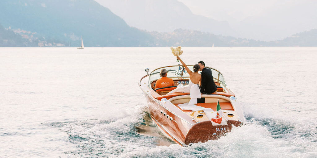 A couple dressed in wedding attire sits on the back of a wooden speedboat, with the bride holding a bouquet aloft as they travel across a lake surrounded by mountains.
