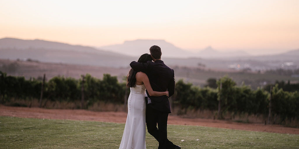 A couple dressed in formal attire stands arm in arm on a lawn, facing a vineyard and distant mountains at sunset.