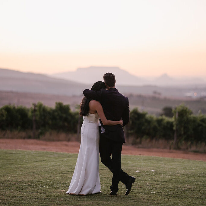 A couple dressed in formal attire stands arm in arm on a lawn, facing a vineyard and distant mountains at sunset.