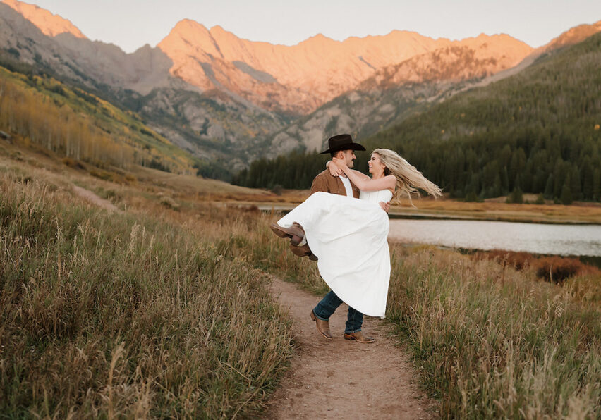 A man in a cowboy hat lifts a woman in a white dress on a grassy path with mountains and a lake in the background at sunset.
