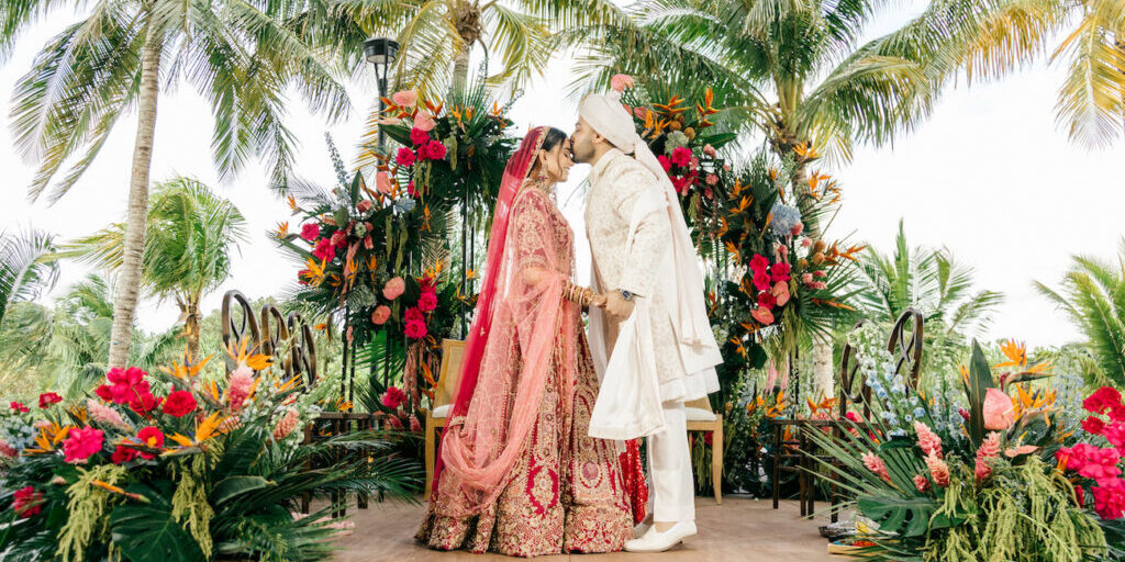 A bride and groom in traditional attire kiss on a decorated outdoor stage surrounded by tropical plants and colorful flowers.