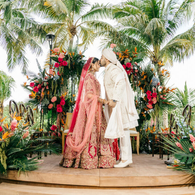 A bride and groom in traditional attire kiss on a decorated outdoor stage surrounded by tropical plants and colorful flowers.