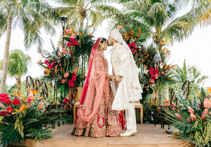 A bride and groom in traditional attire kiss on a decorated outdoor stage surrounded by tropical plants and colorful flowers.