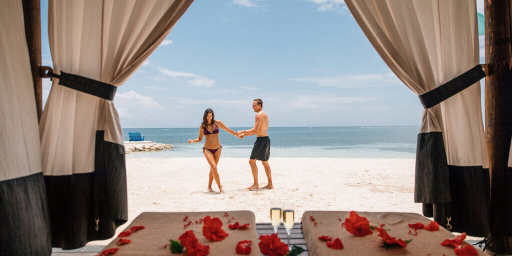 A couple holds hands on a sandy beach, viewed from a cabana with curtains. Red flowers and towels are on a table in the foreground.