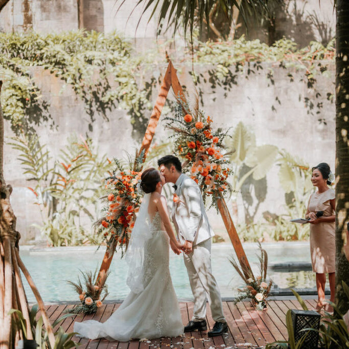 Bride and groom share a kiss at their outdoor wedding ceremony, surrounded by tropical plants and standing in front of a triangular floral arch. Guests watch and capture the moment.