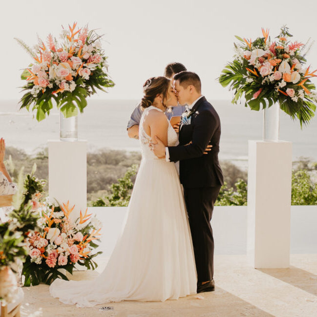 A couple shares a kiss at their outdoor wedding ceremony. They stand between two large floral arrangements, with a scenic ocean view in the background. Guests are seated on either side, capturing and watching the moment. The sun shines brightly, illuminating the scene.