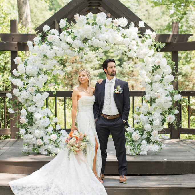 A bride and groom stand side by side on a wooden platform, in front of a large circular floral arrangement outdoors.