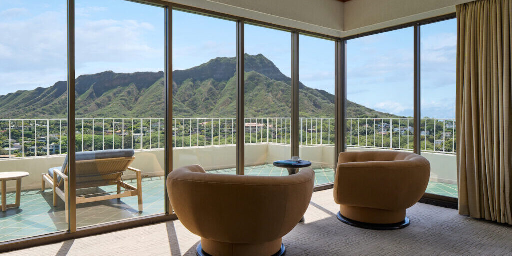 Hotel room with two curved chairs facing large windows, offering a view of a mountain landscape and a balcony with a lounge chair and small table.