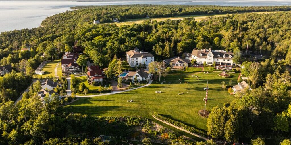 Aerial view of several large houses and open green lawns surrounded by dense forest near a body of water.
