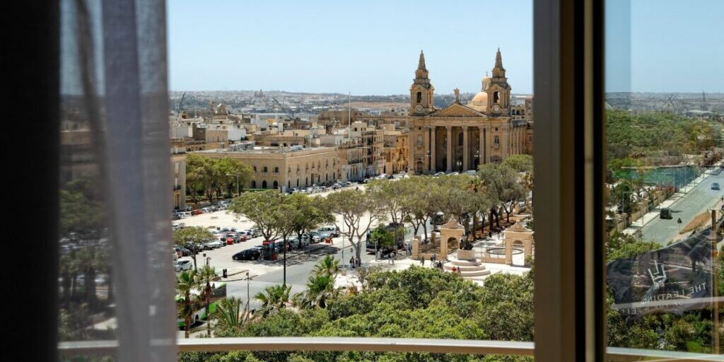 View through a window overlooking a city square with trees, parked cars, and a large historic church with two bell towers, under a clear sky.