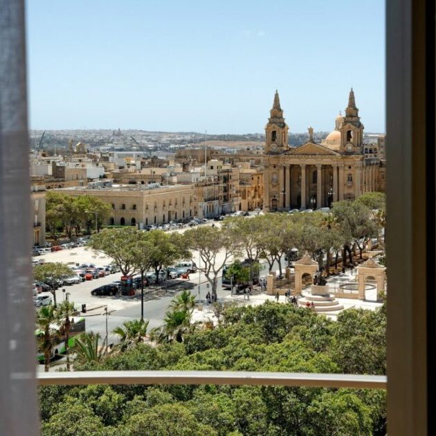 View through a window overlooking a city square with trees, parked cars, and a large historic church with two bell towers, under a clear sky.