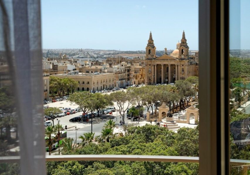 View through a window overlooking a city square with trees, parked cars, and a large historic church with two bell towers, under a clear sky.
