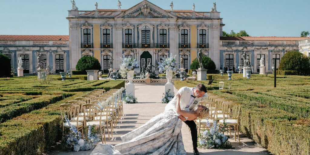 A couple poses in a dip at an outdoor wedding ceremony, surrounded by gold chairs and floral arrangements, in front of an ornate historic building.