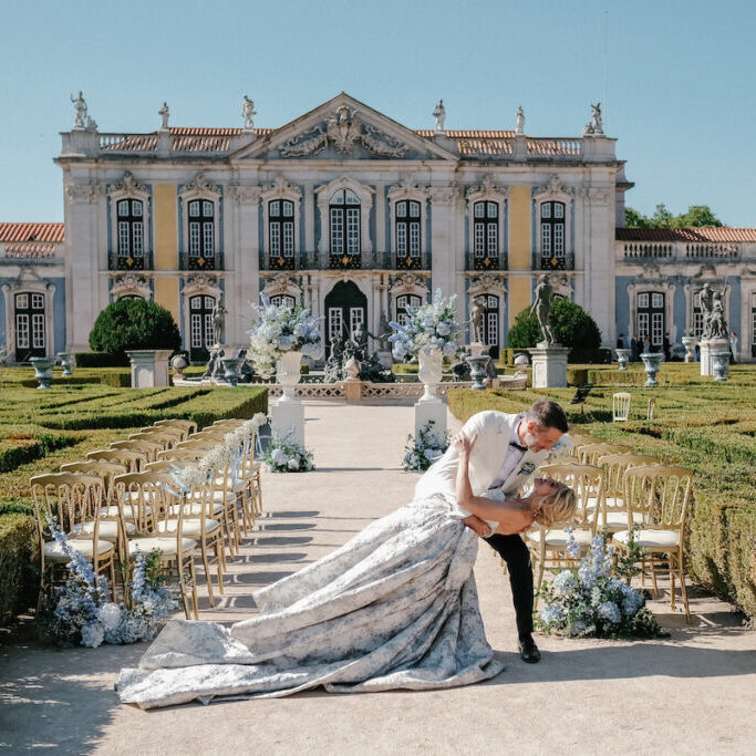 A couple poses in a dip at an outdoor wedding ceremony, surrounded by gold chairs and floral arrangements, in front of an ornate historic building.