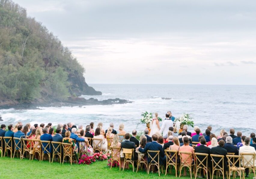 A wedding ceremony takes place outdoors near the ocean, with guests seated on wooden chairs. The background features a rocky coastline and overcast sky.