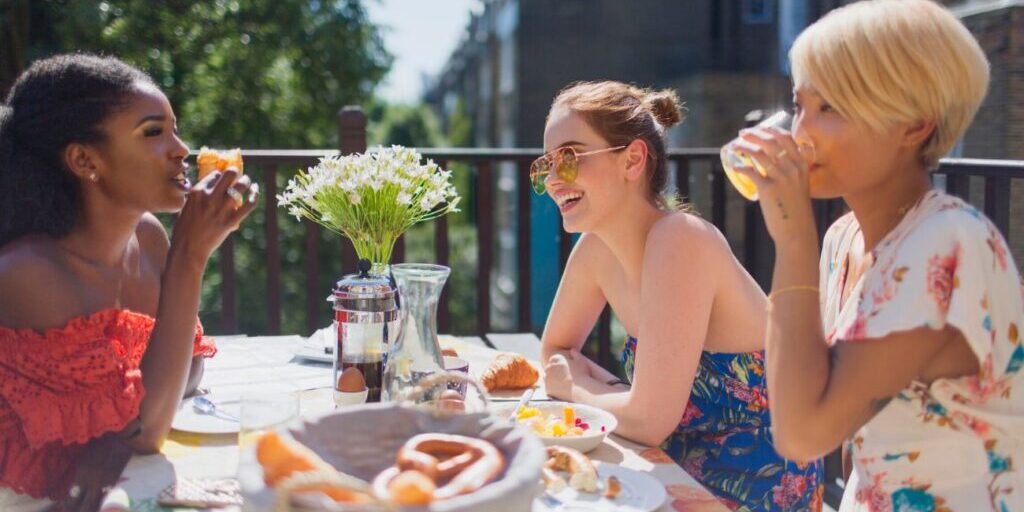 Three people sit at an outdoor table with food and drinks, talking and smiling. A vase of white flowers is in the center of the table.