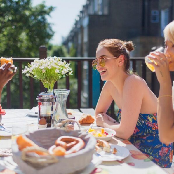 Three people sit at an outdoor table with food and drinks, talking and smiling. A vase of white flowers is in the center of the table.